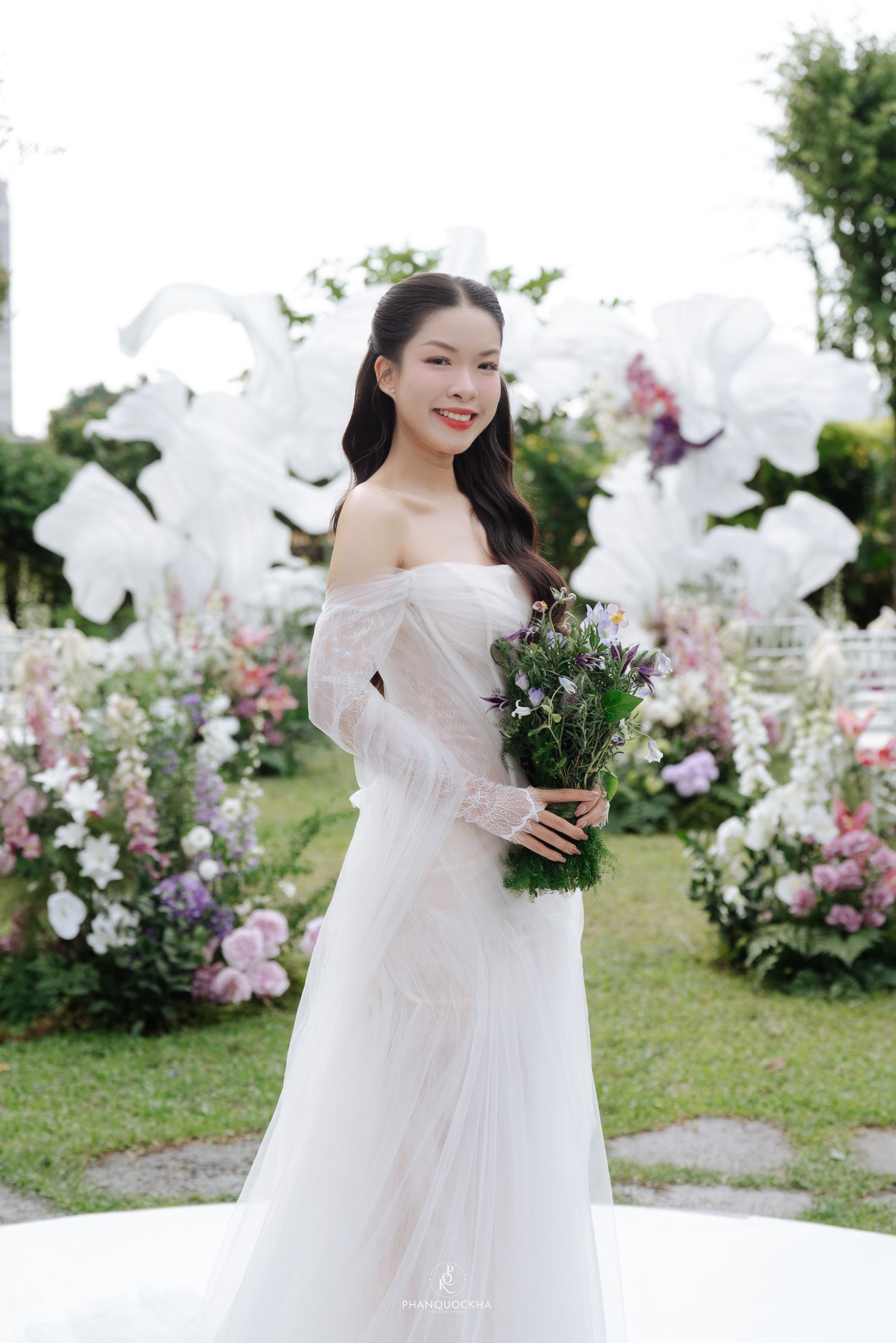 Bride in off-shoulder white wedding gown holding purple bouquet at outdoor floral ceremony.
