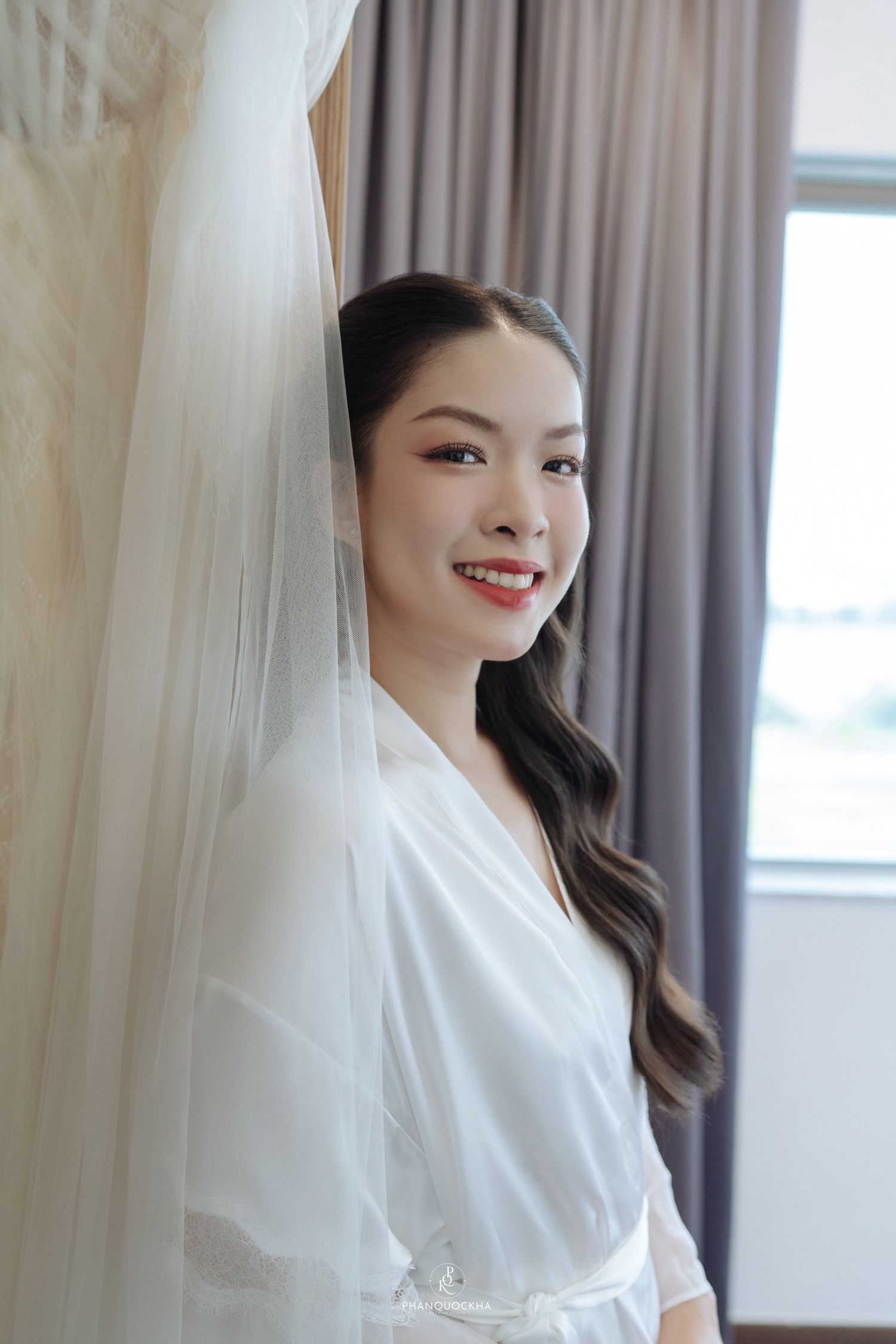 Smiling Asian bride in white robe posing beside her wedding veil before the ceremony.