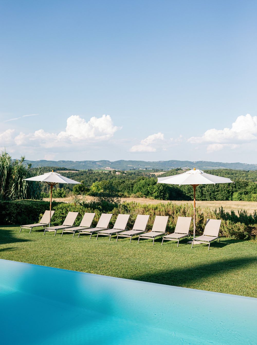 Scenic infinity pool with lounge chairs and umbrellas overlooking rolling green hills in the Italian countryside at Borgo Stomennano.