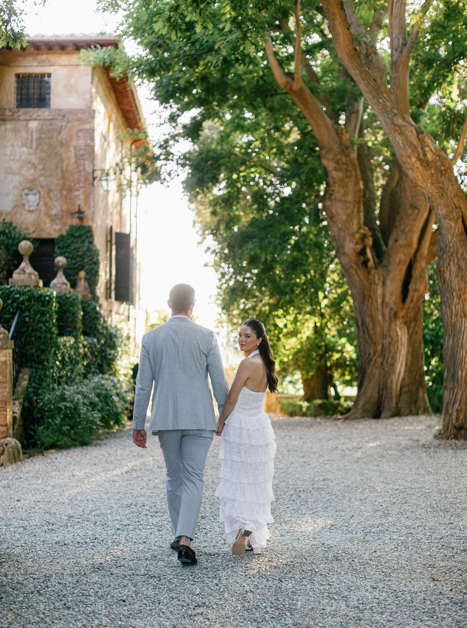 A couple walks hand in hand on a stone path under lush green trees at a rustic Borgo Stomennano.