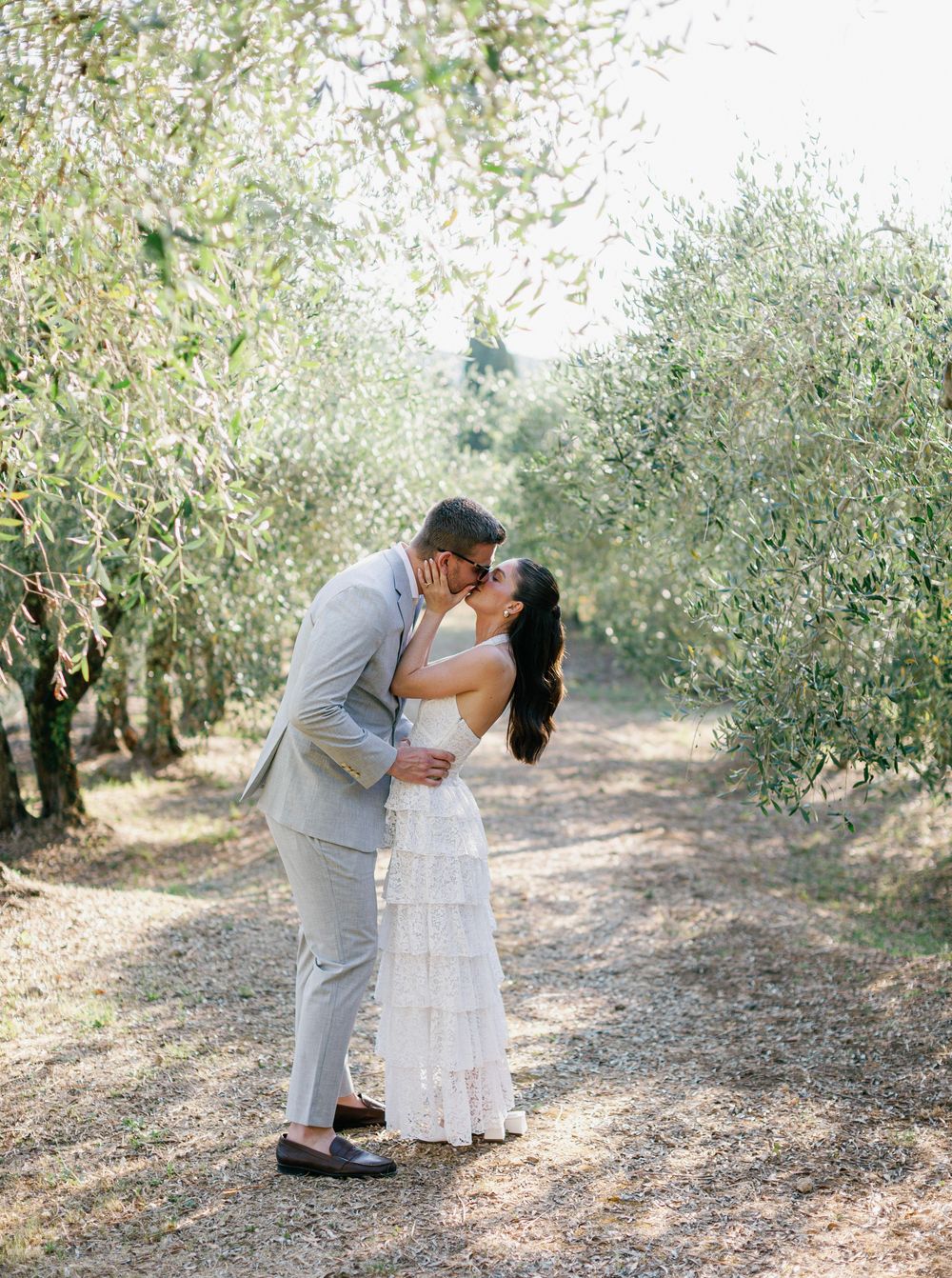 A couple shares romantic kisses in an olive grove wearing a light gray suit and white dress during a sunny outdoor celebration in Tuscany.
