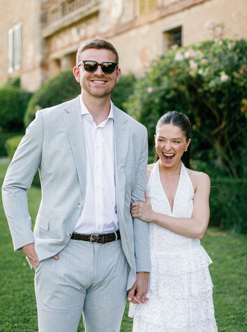 A couple wearing white and gray outfits share playful candid moments in an outdoor garden setting at Borgo Stomennano.