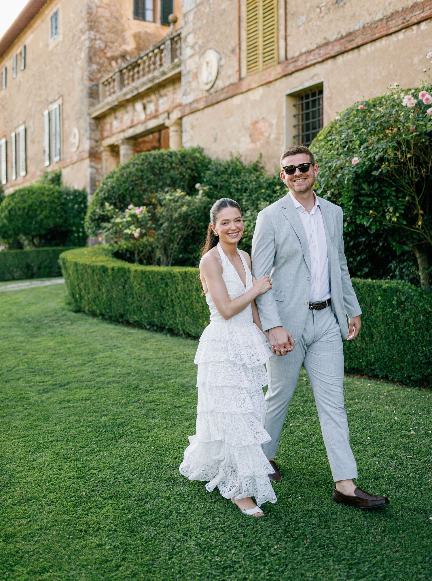 A couple in wedding attire shares a romantic walk and laugh together on a lush green lawn by an old villa Borgo Stomennano.