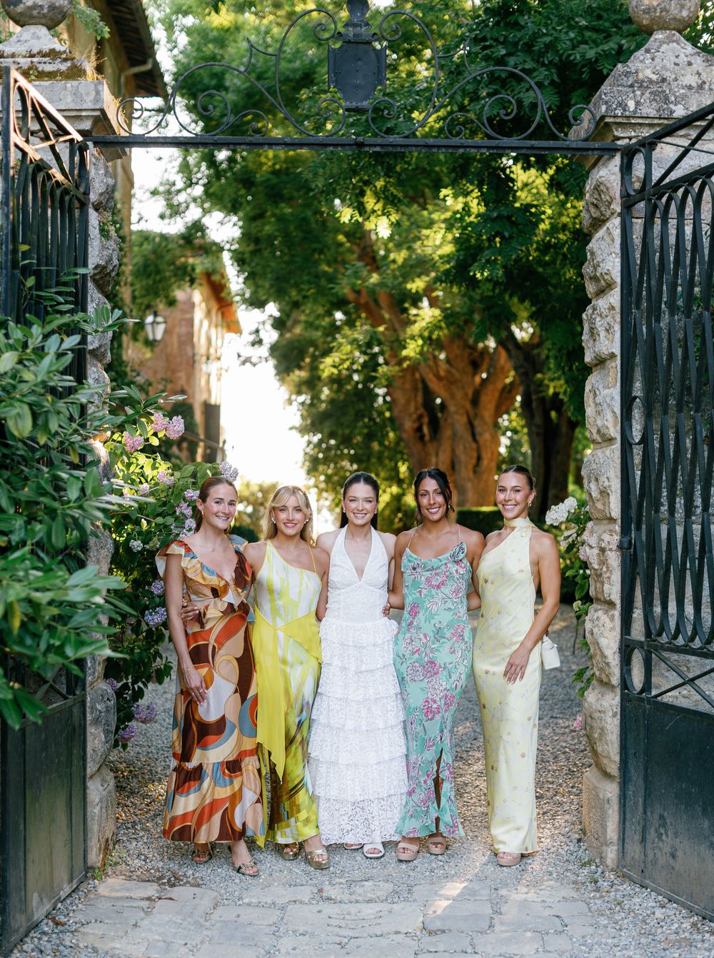 Group in pastel and floral dresses poses among lush garden foliage by decorative iron gates.