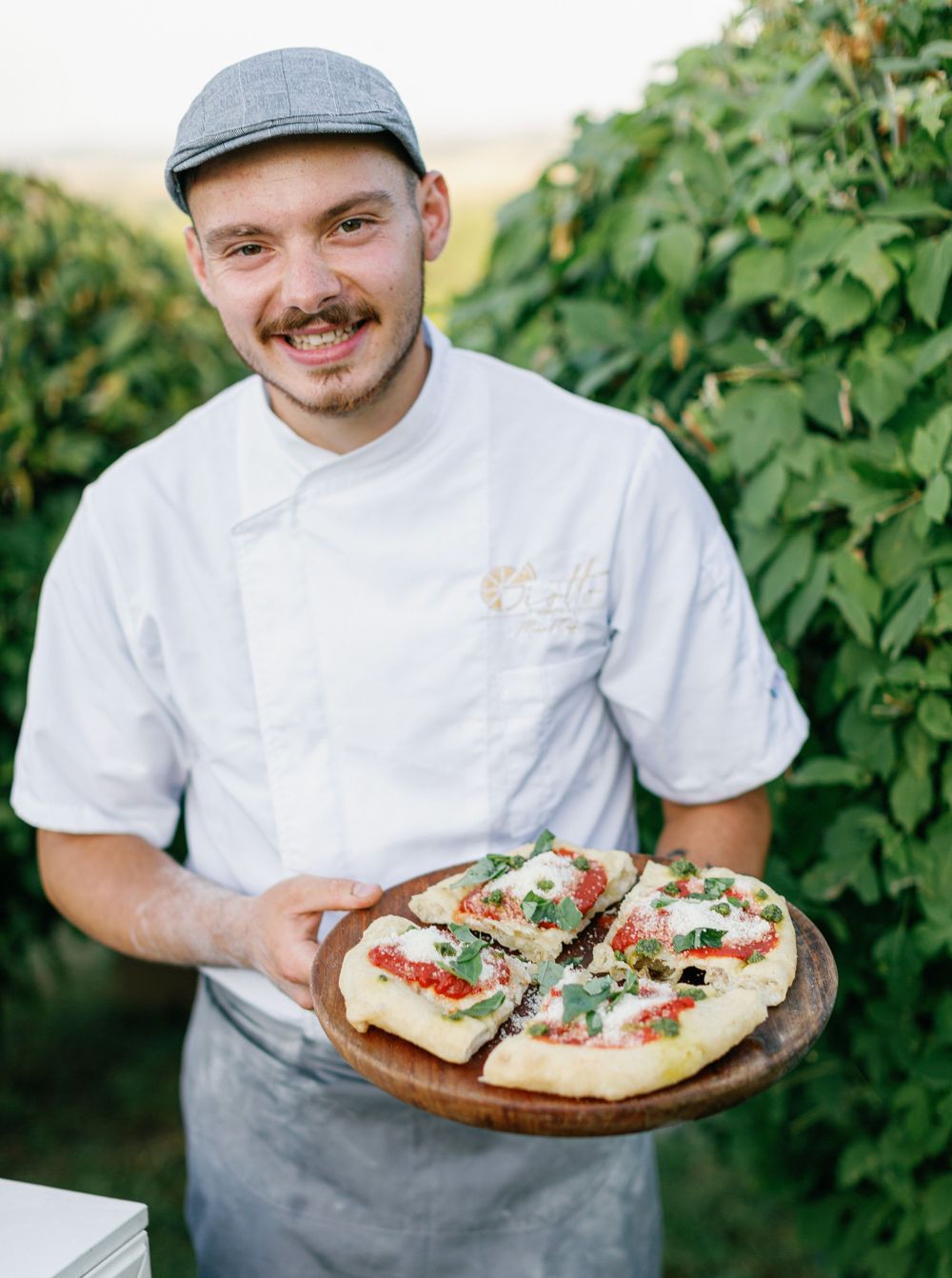 Chef smiles while holding a wooden cutting board with fresh pizza at outdoor garden restaurant.