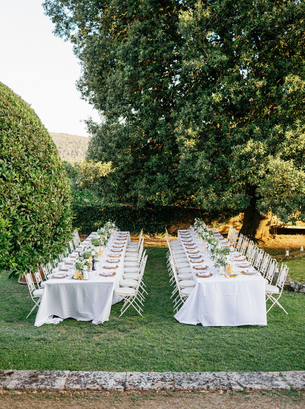 Elegant outdoor wedding reception setup with long white dining tables arranged on a grassy lawn under lush green trees.