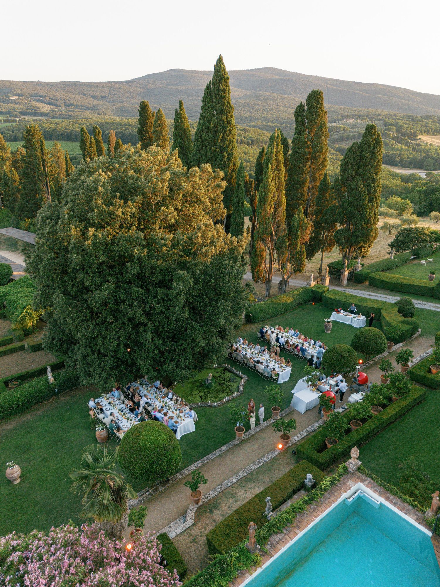 Aerial view of an elegant outdoor wedding reception setup with white tables amidst towering cypress trees in Tuscany at Borgo Stomennano.