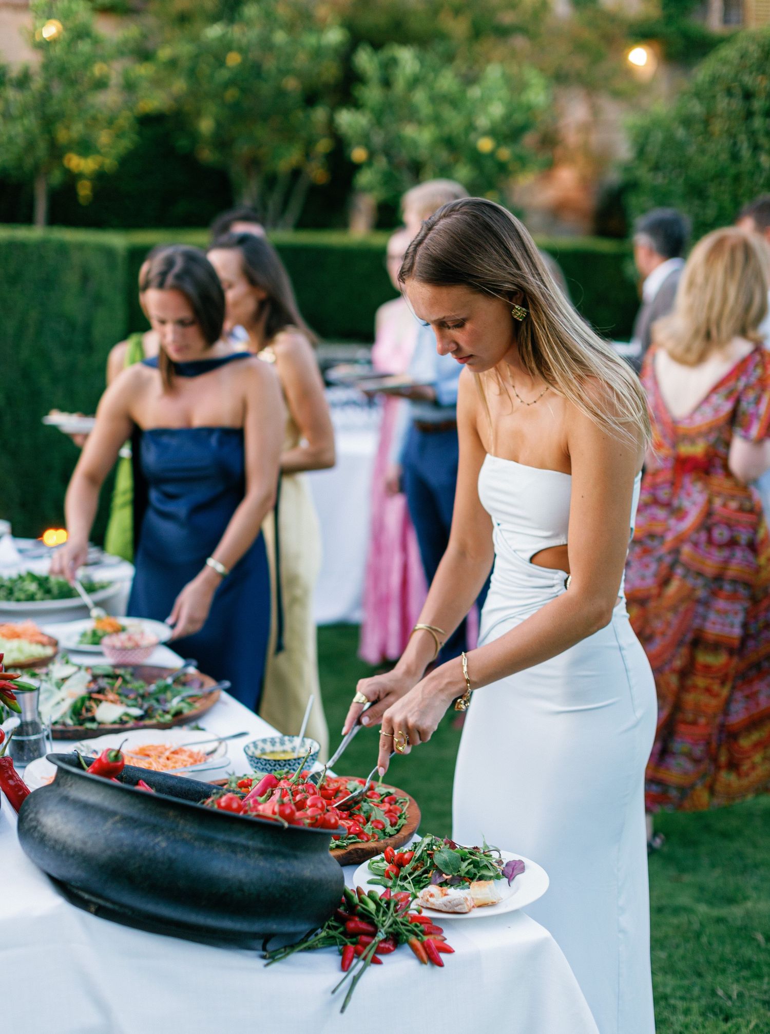 A bride in white dress serving food from an outdoor buffet table at a garden wedding reception.