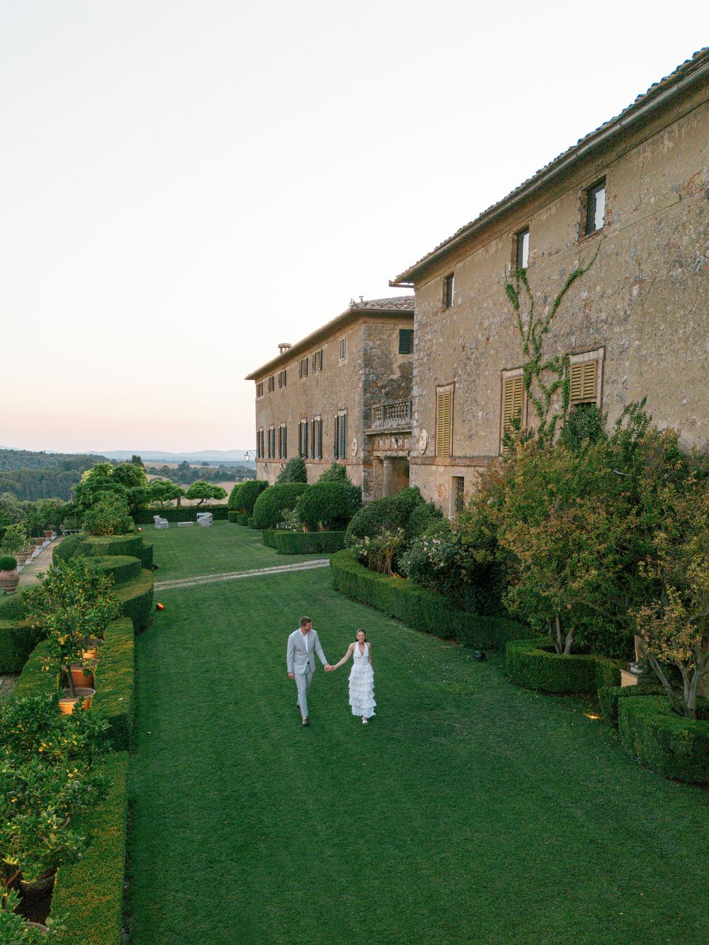 Two people stroll along a manicured lawn garden path beside a grand Italian villa Borgo Stomennano during sunset.