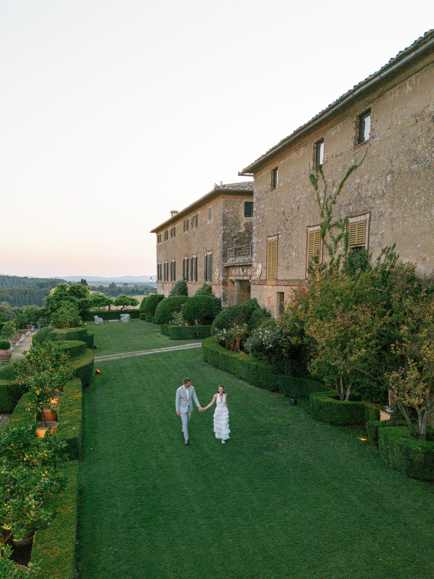Two people stroll along a manicured lawn garden path beside a grand Italian villa Borgo Stomennano during sunset.