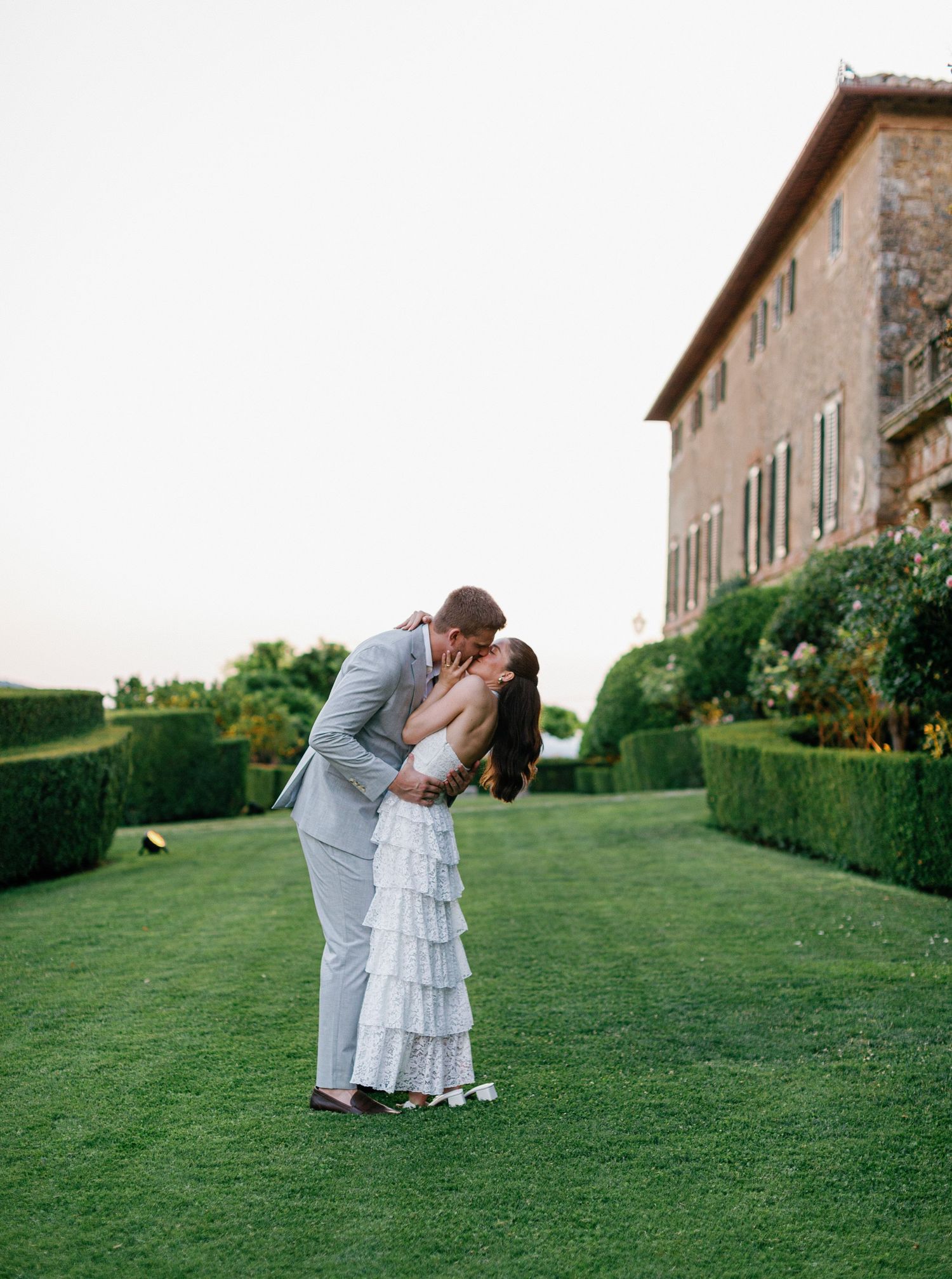 A romantic couple shares a tender moment on a manicured lawn in front of an elegant Italian villa Borgo Stomennano.