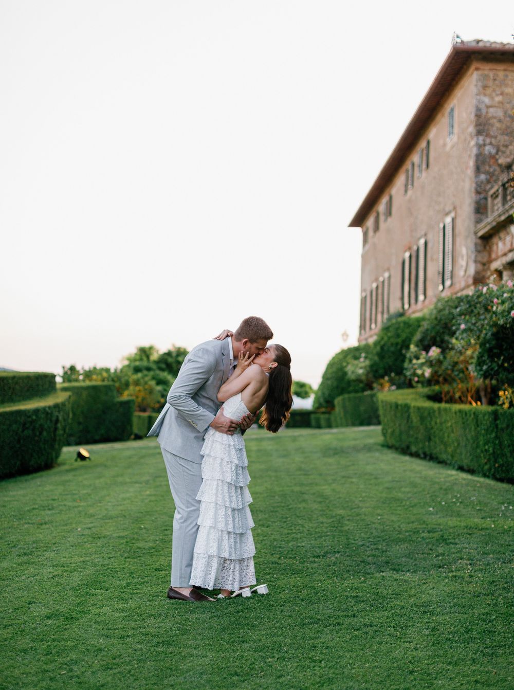 A romantic couple shares a tender moment on a manicured lawn in front of an elegant Italian villa Borgo Stomennano.