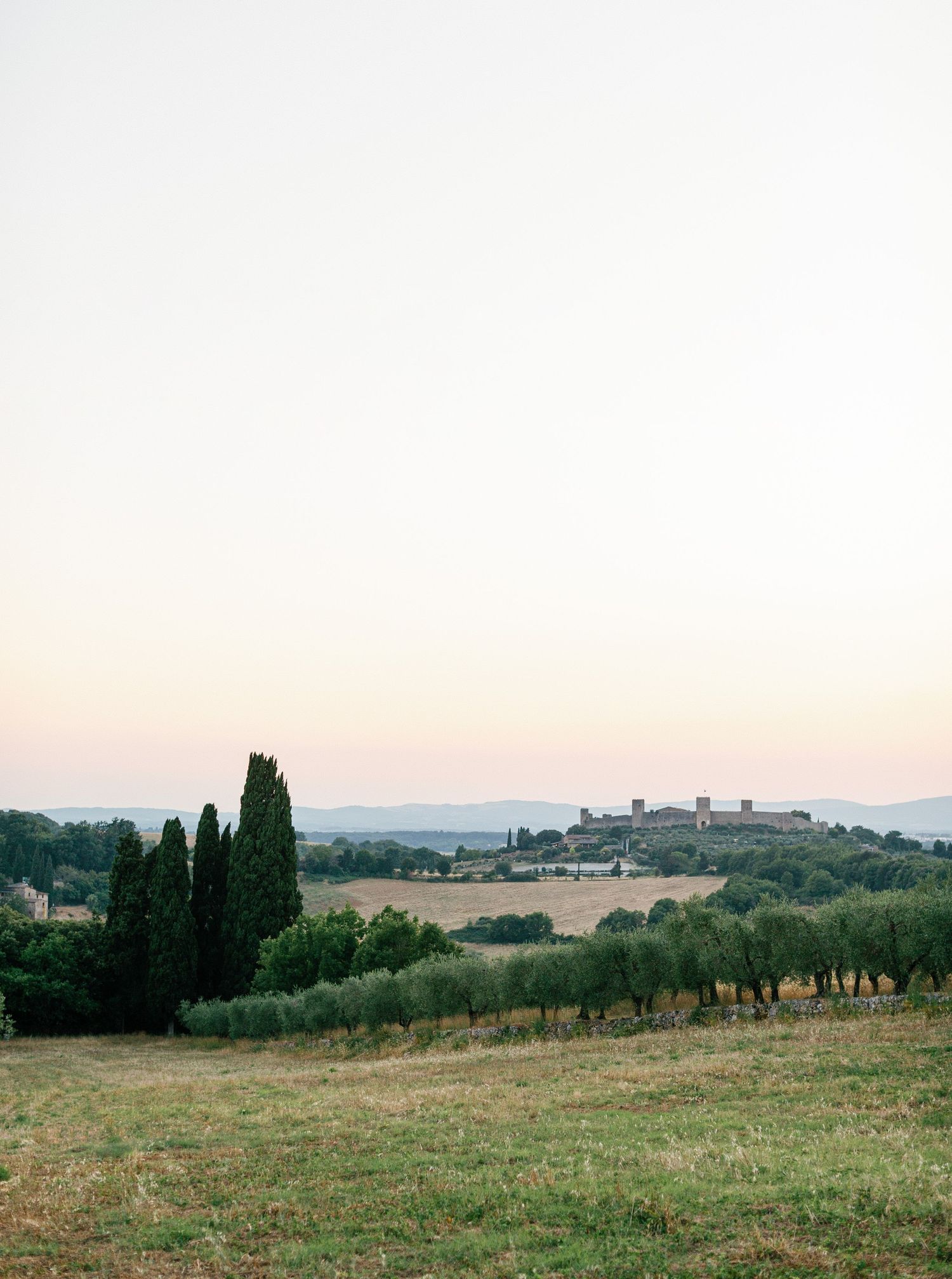 Panoramic view of rolling Tuscan countryside with cypress trees and hilltop village silhouetted against sunset sky.