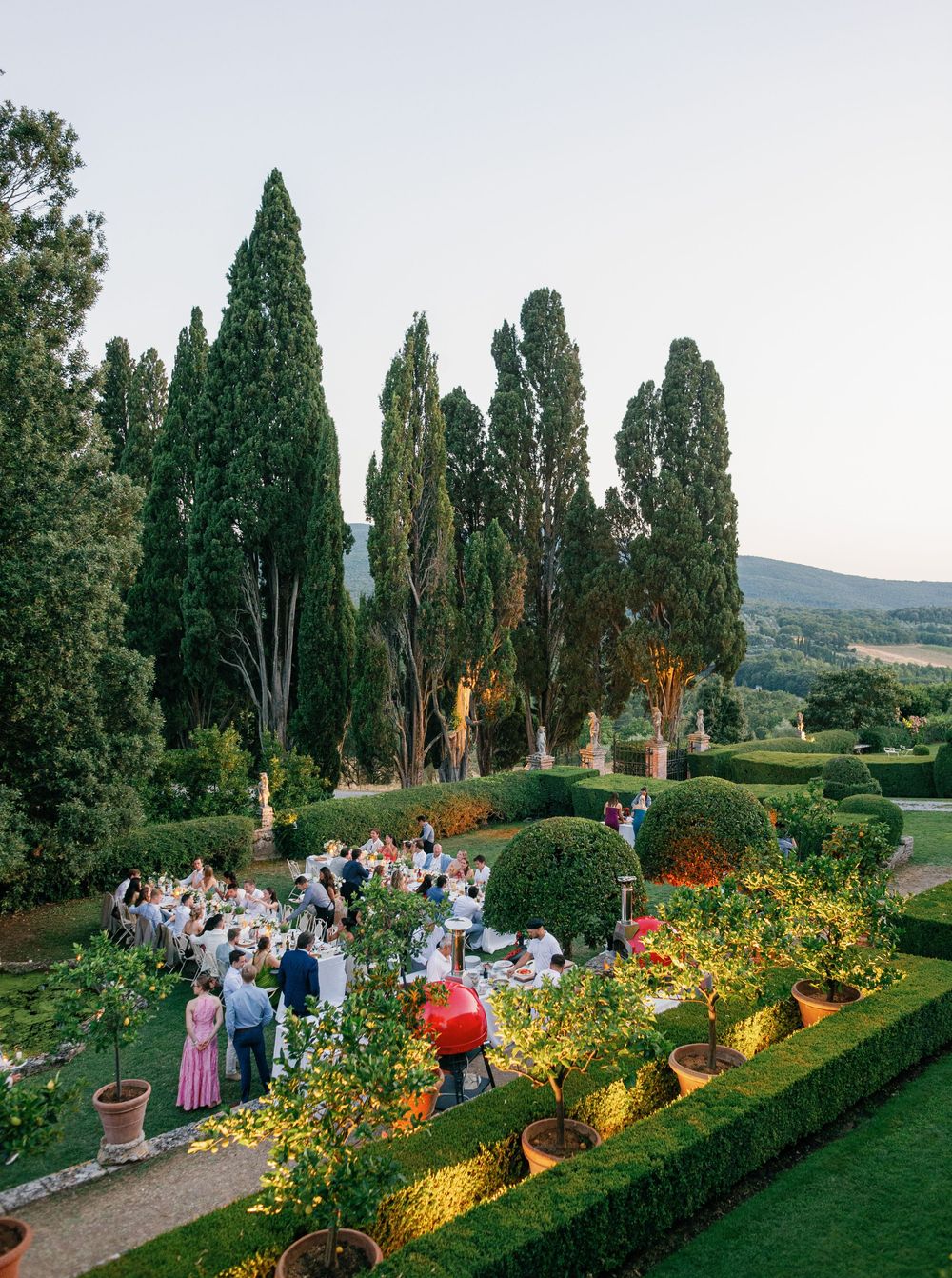 Garden party guests enjoying an evening outdoor celebration along illuminated cypress trees and flower beds in Tuscany at Borgo Stomennano.
