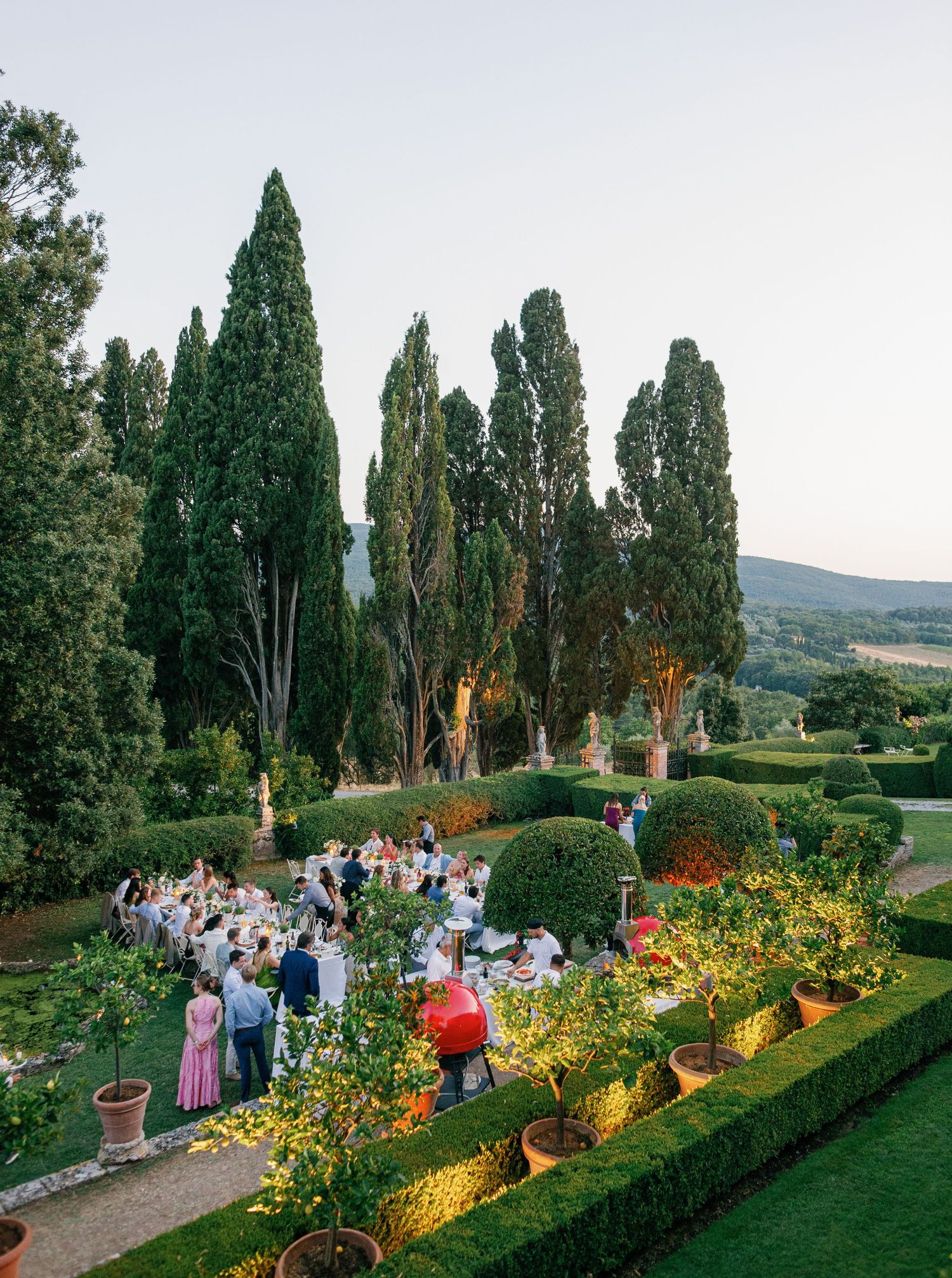 Garden party guests enjoying an evening outdoor celebration along illuminated cypress trees and flower beds in Tuscany at Borgo Stomennano.