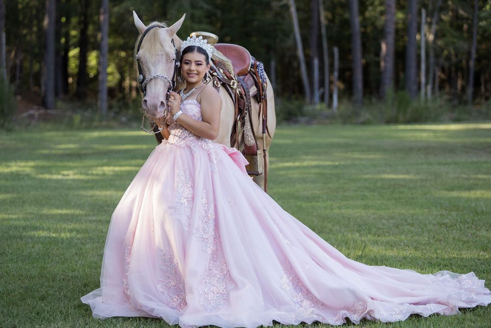 Someone wearing a pink lace ballgown poses in a sunlit outdoor garden setting while holding flowers and smiling.