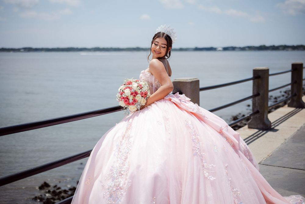 A person in a flowing pink ballgown poses elegantly by a waterfront railing during a photo session.