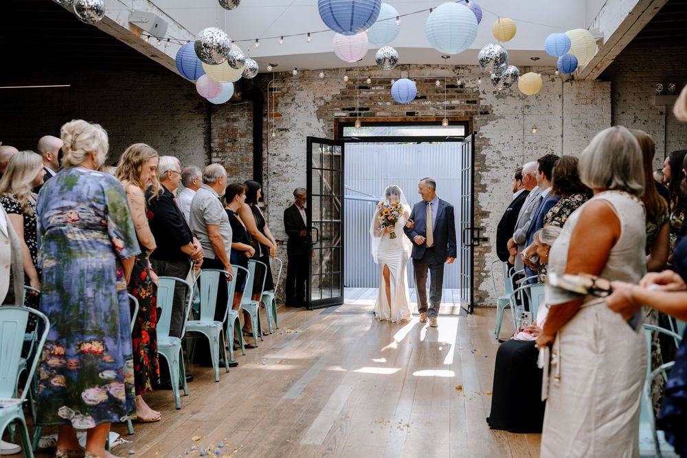 Wedding guests sit on white chairs in two sections during an indoor ceremony decorated with hanging paper lanterns.