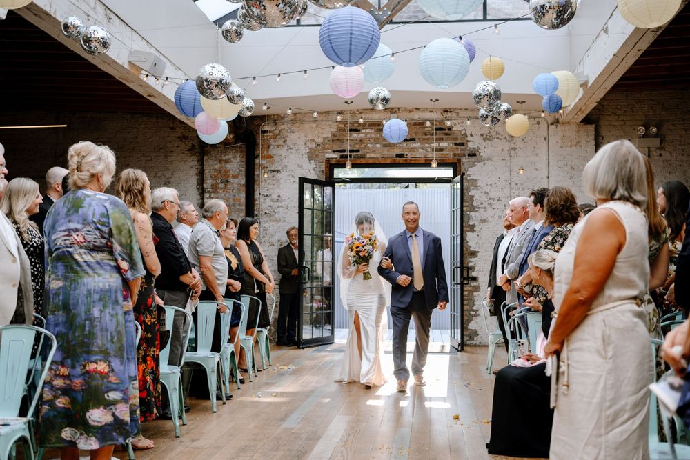 Wedding guests gather in an industrial-style venue decorated with blue paper lanterns and exposed brick walls.