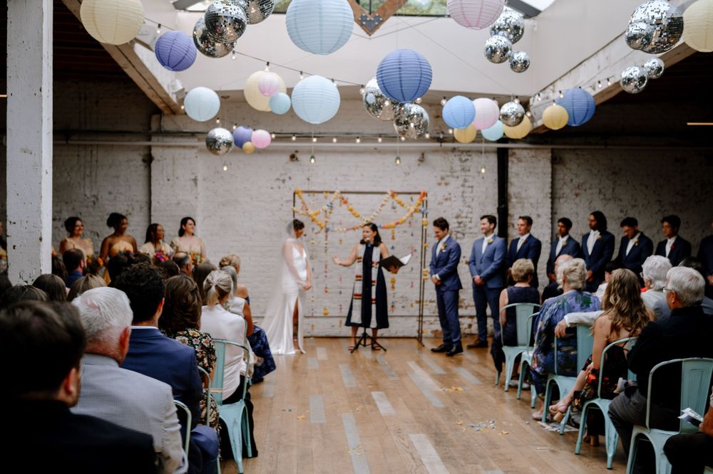 Wedding guests seated in turquoise chairs with pastel balloon decorations overhead in an urban venue.