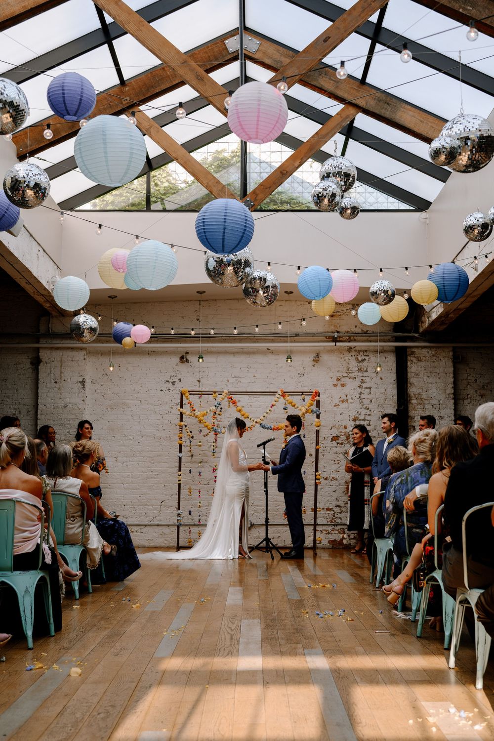 Wedding ceremony in a rustic indoor venue with glass ceiling and blue and pink paper lanterns decorations.