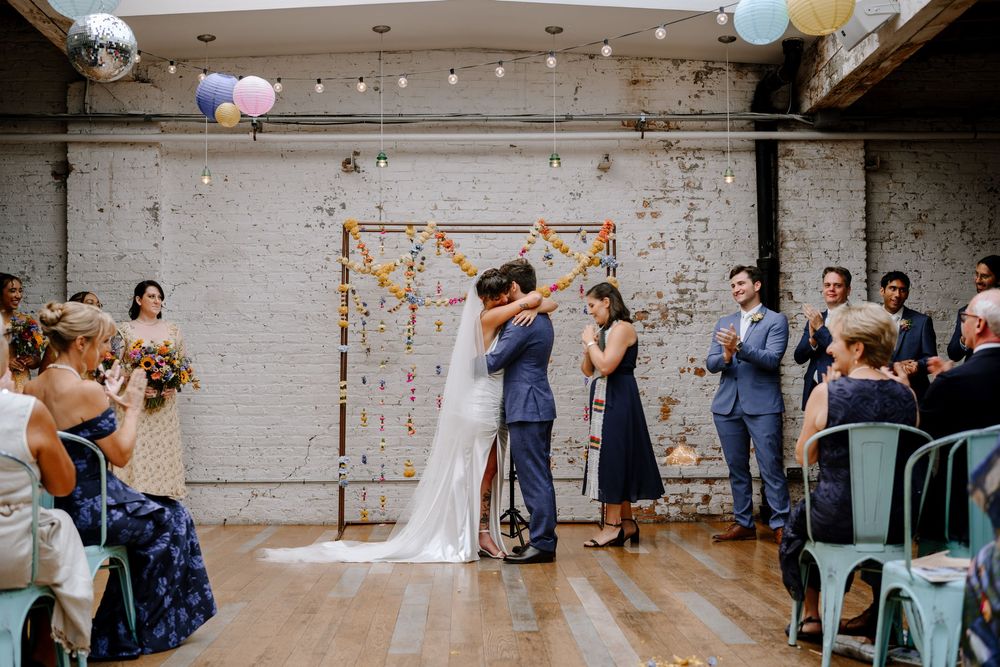 Wedding guests seated at long tables in an industrial space decorated with hanging paper lanterns and floral accents.