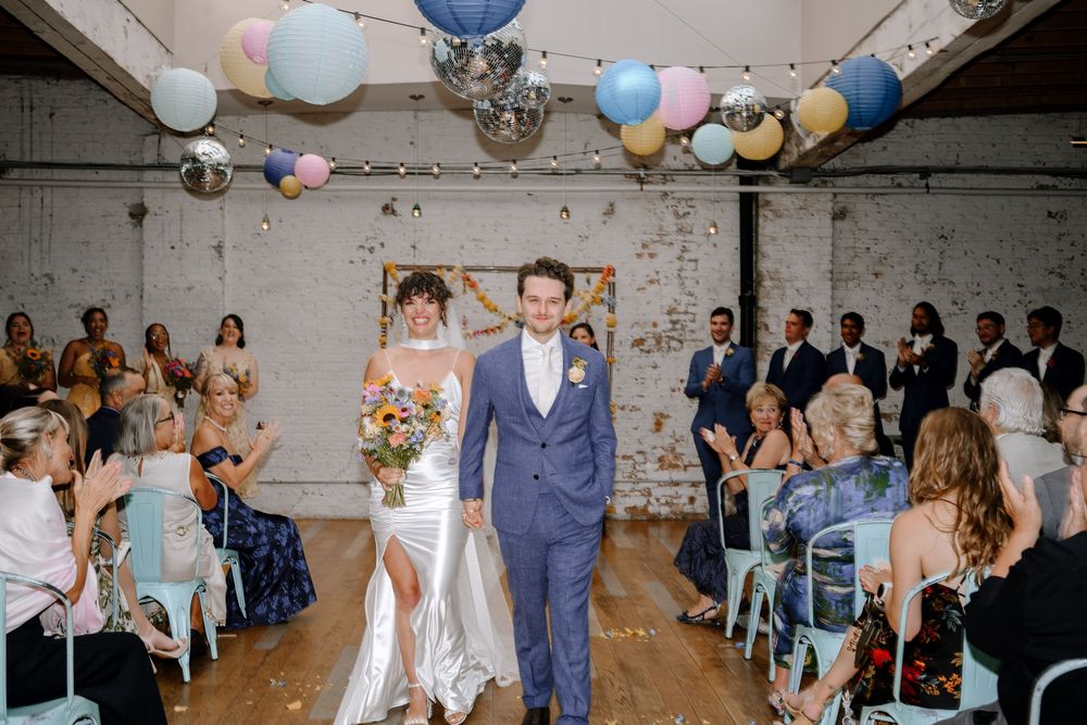 Indoor wedding ceremony with white brick walls, glass ceiling, and pastel colored lantern decorations.