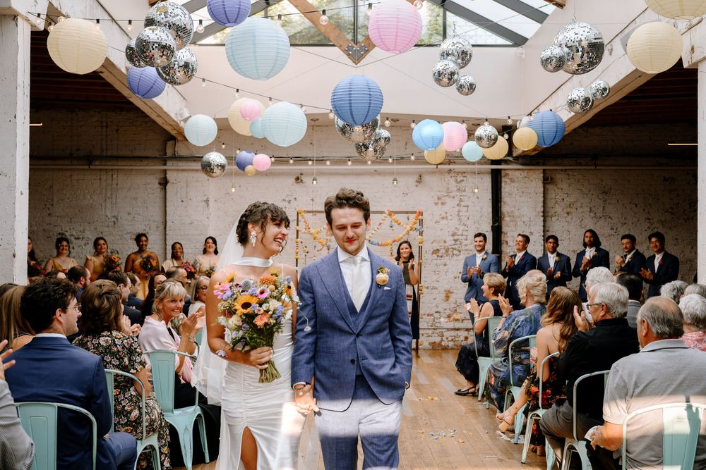 Wedding ceremony set in an industrial venue with exposed brick walls and colorful hanging paper lanterns.
