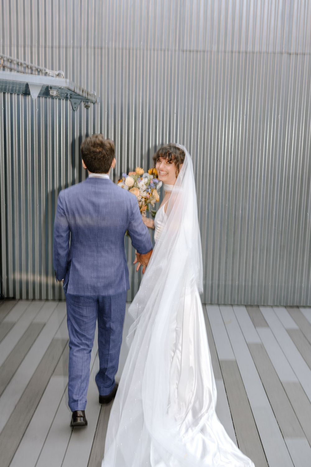A bride and groom walk through an industrial hallway with corrugated metal walls and wooden flooring.
