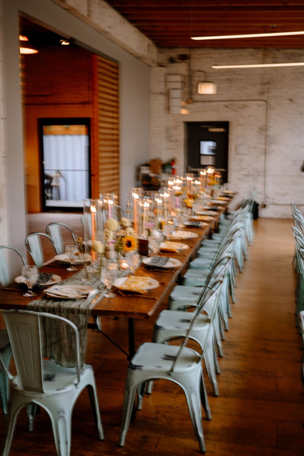 Long dining tables with mint green metal chairs set for an elegant event in an industrial-style space with string lighting.