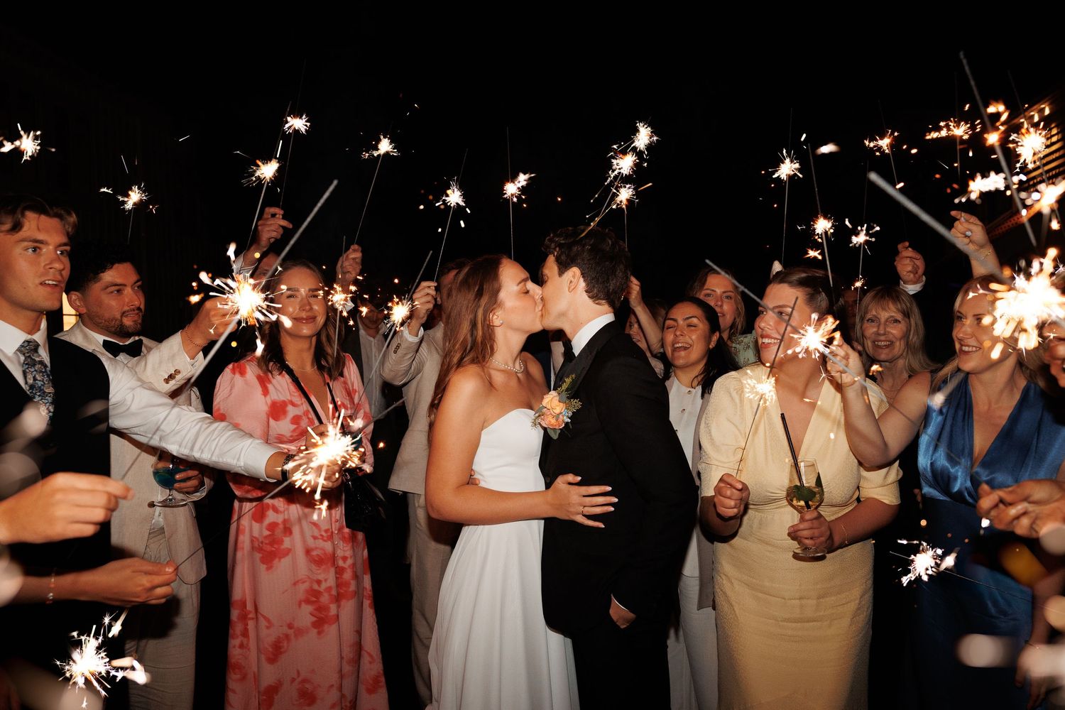 Wedding party guests holding sparklers in the dark creating a magical nighttime celebration atmosphere.