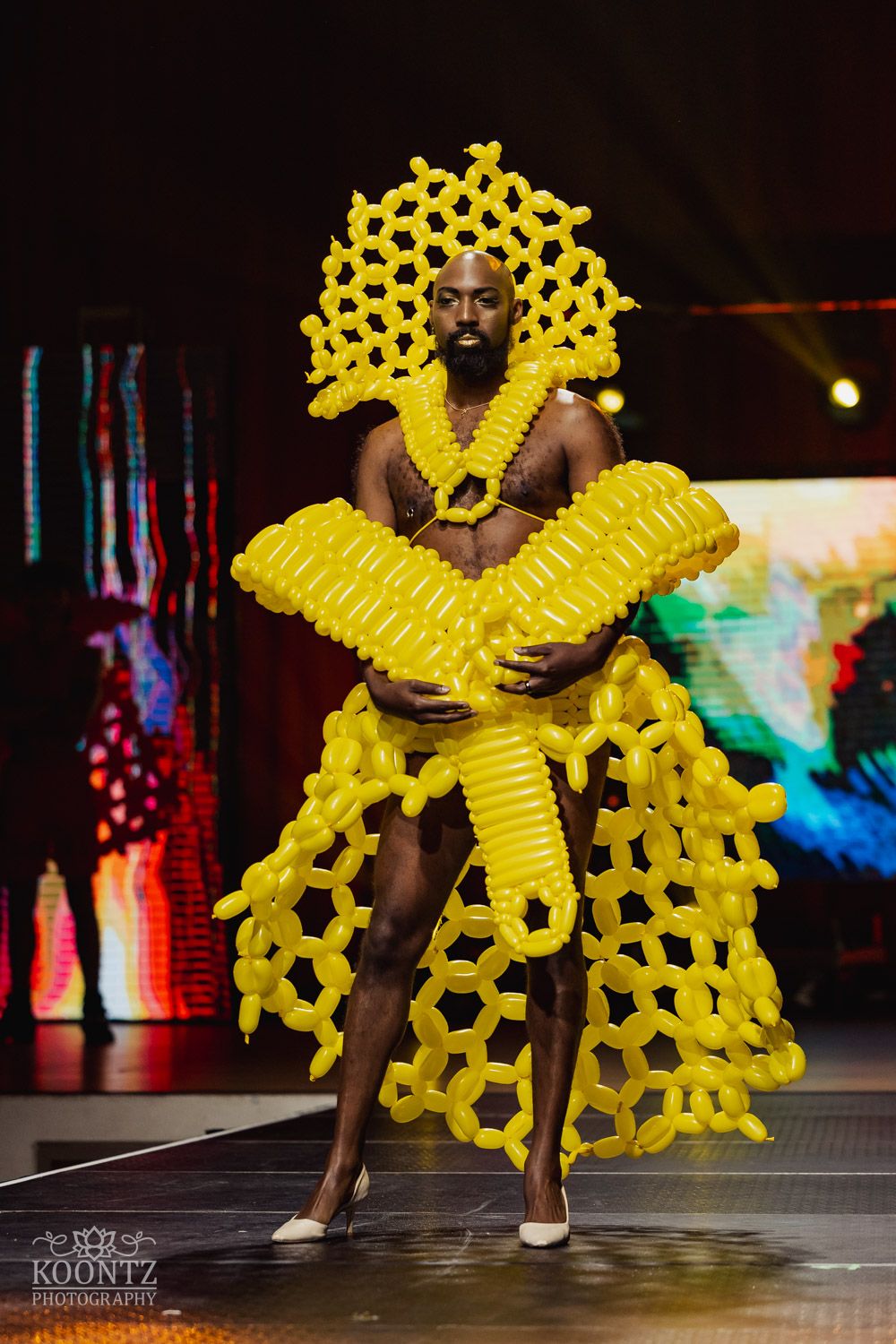 Performer in avant-garde yellow balloon costume strutting dramatically on dark runway stage with colorful lighting.