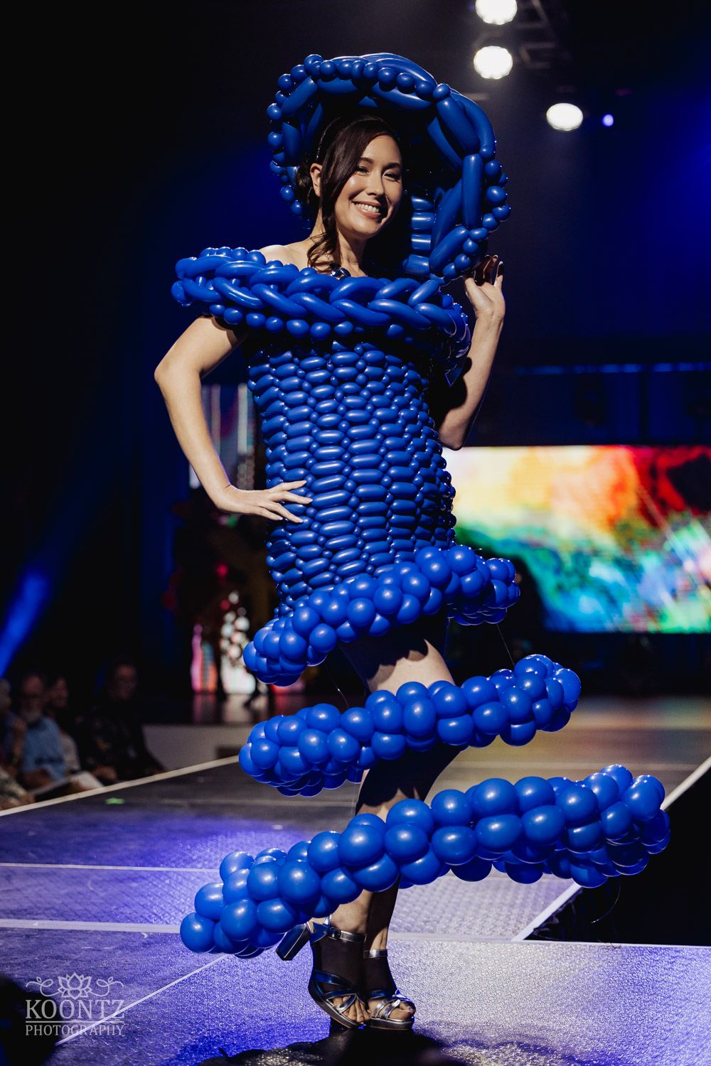 Performance artist in a sculptural costume made entirely of bright blue balloons on a dark stage.