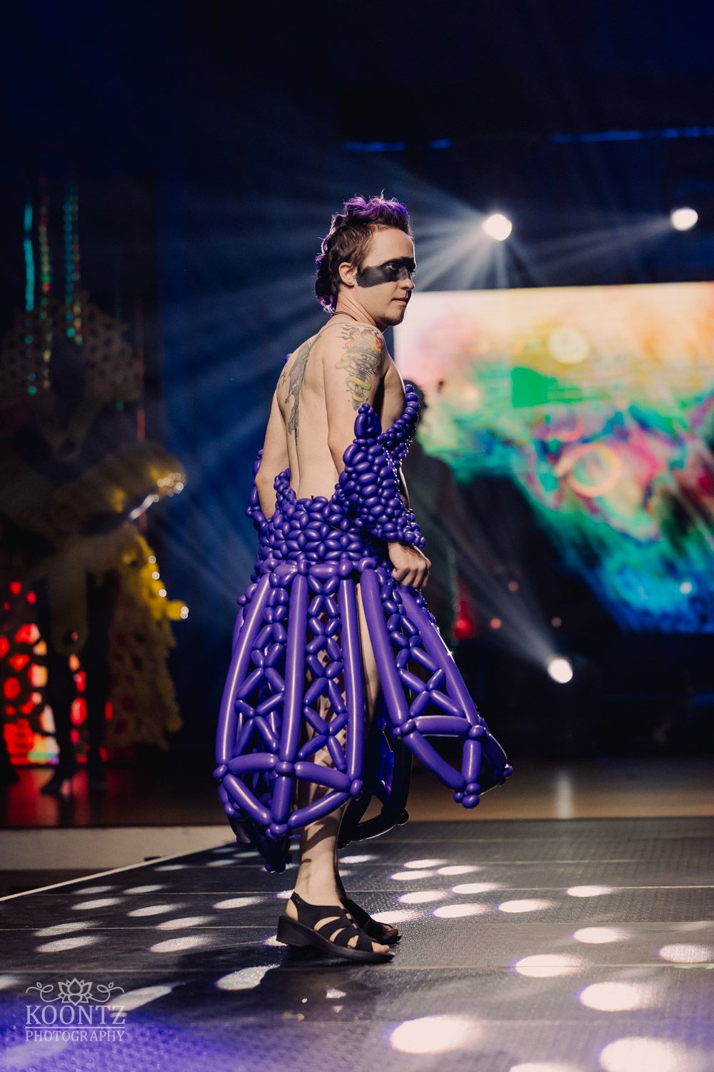 Dramatic fashion show featuring deep purple balloon-style dress on illuminated catwalk with spotlight effects.