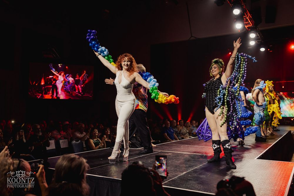 Performers take final bows on stage during colorful fashion show finale with dramatic lighting.