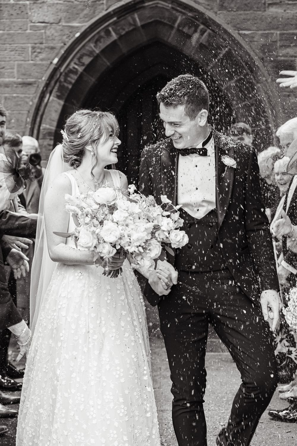 A wedding couple and guests celebrate with joyful confetti throwing outside a stone church entrance.