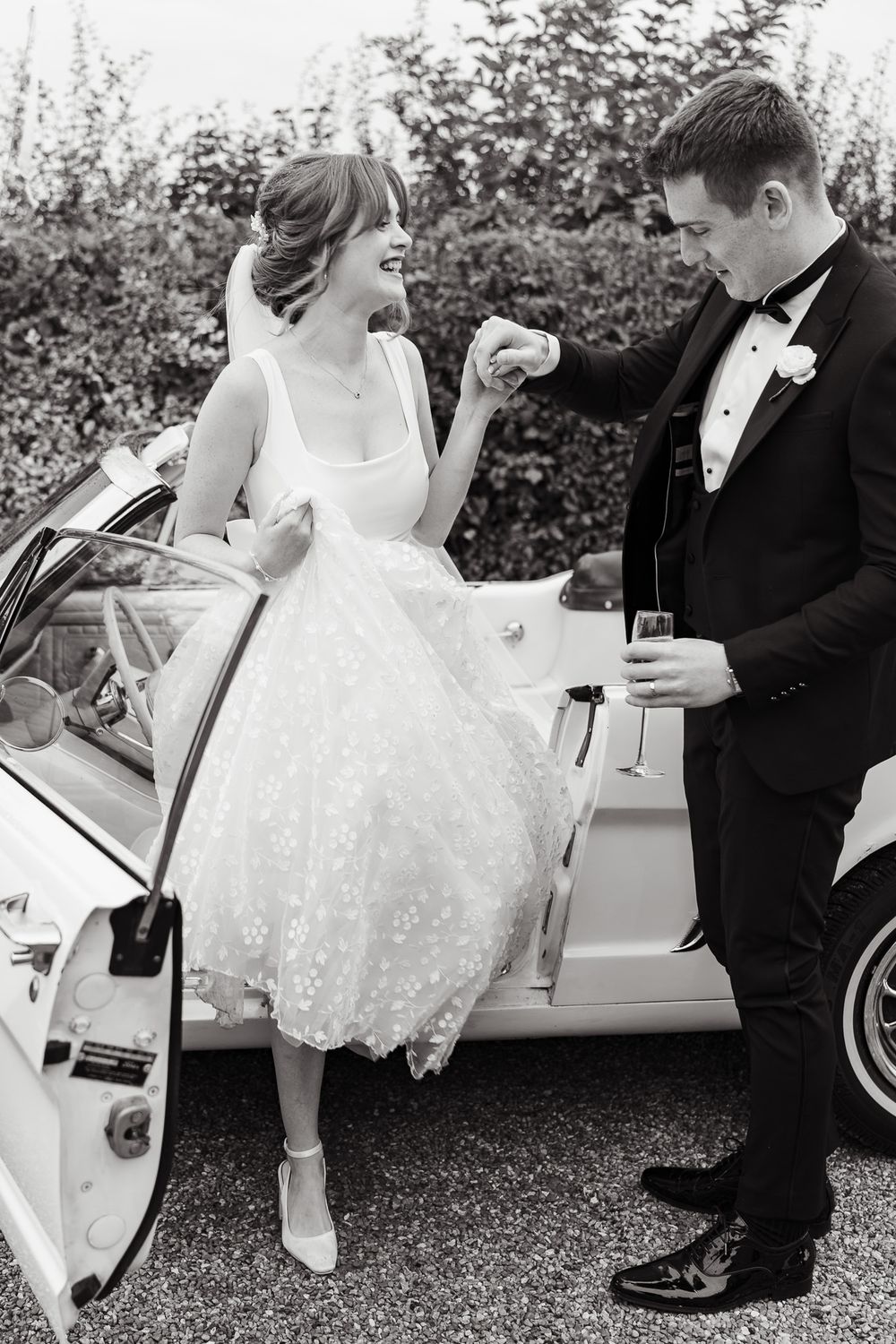 Nostalgic black and white wedding photo showing couple in vintage attire sharing champagne by classic car outdoors.