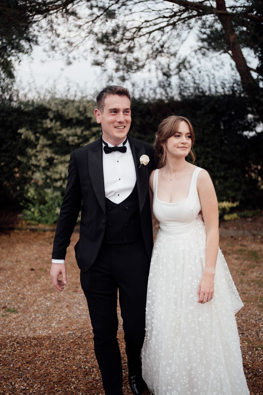 A wedding couple walks outdoors through a wooded area, with the bride in white and groom in classic black tuxedo.