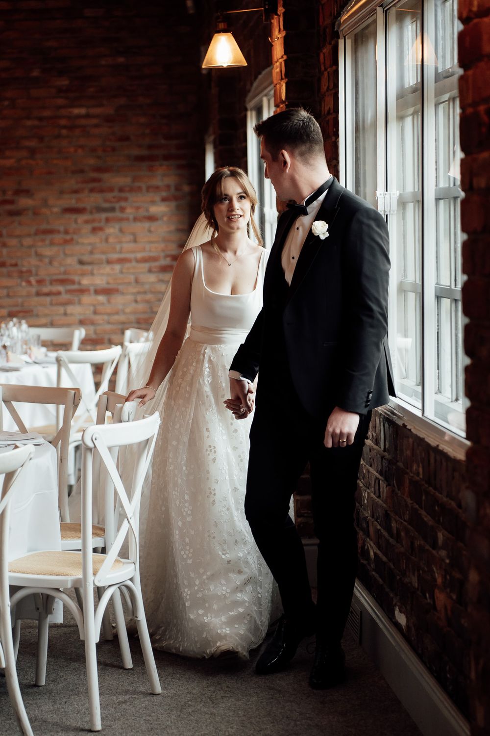 A bride in white lace dress and groom in black suit stand in an elegant rustic brick venue with white chairs near windows.