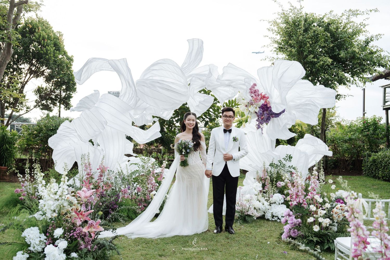Bride and groom standing before white floral arch with pink flowers at outdoor garden wedding.