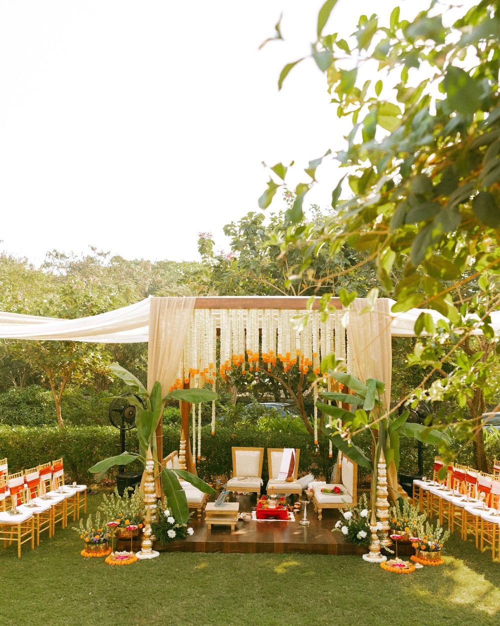 Elegant outdoor wedding setup with wooden furniture and orange floral decorations in a garden setting.