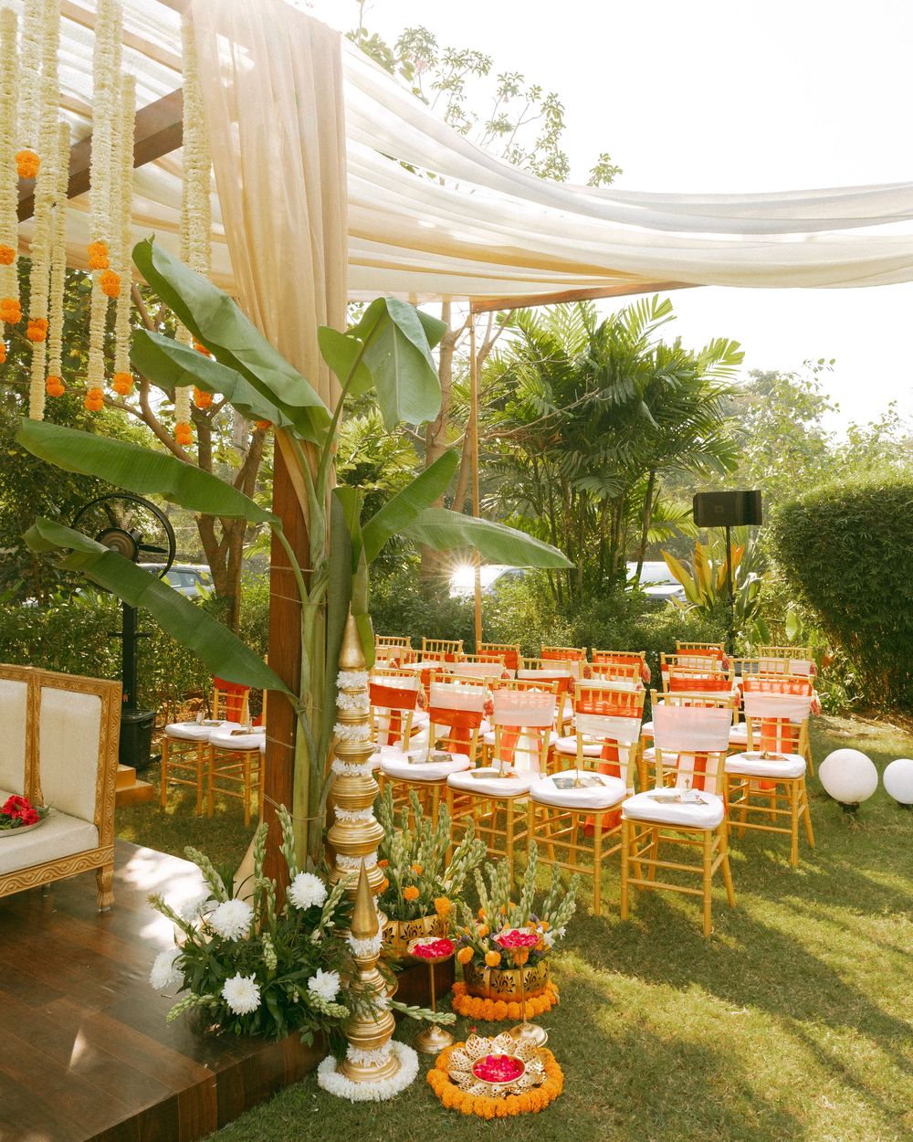Wedding reception area decorated with white drapes, orange flowers and wooden chairs under natural light.