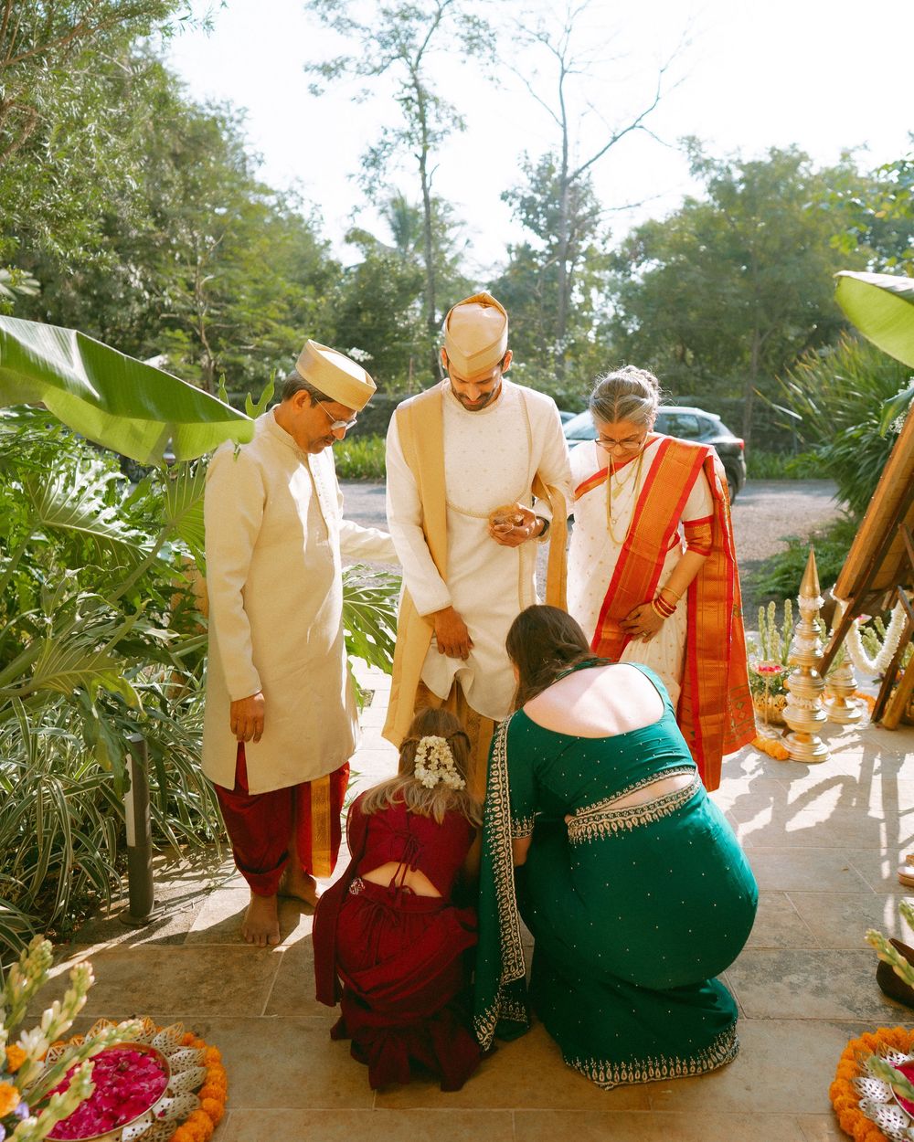 Group wearing traditional Indian attire including saris and kurtas gather outdoors under trees for a special occasion.