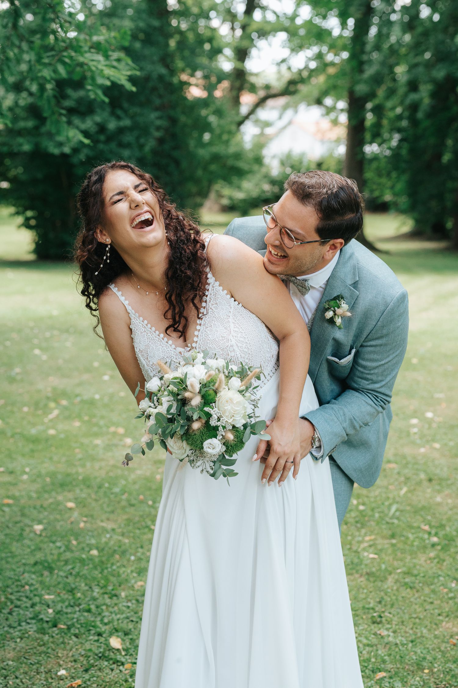 A joyful wedding couple laughs together in a green garden setting while wearing white dress and grey suit.
