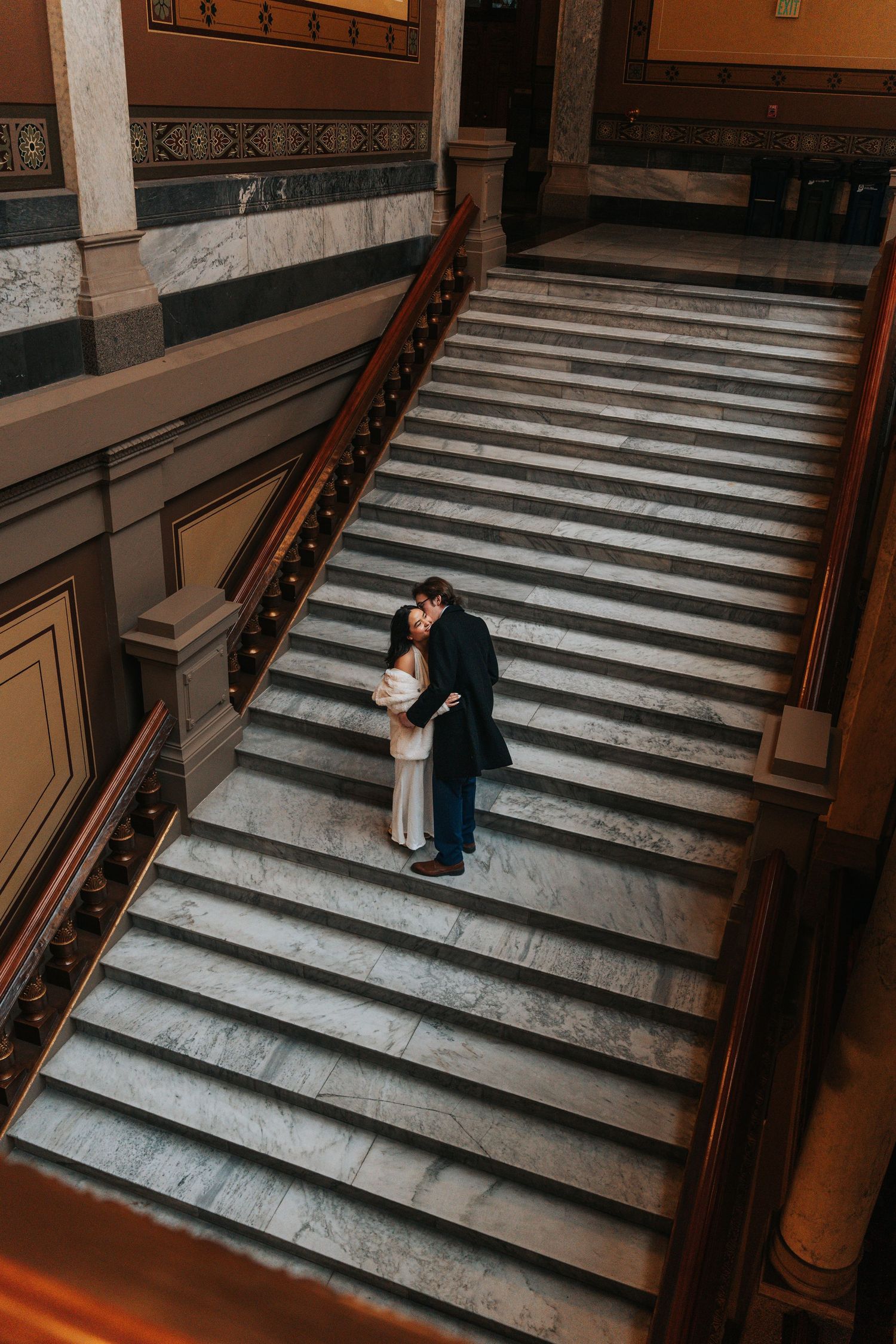 Couple embracing on grand marble staircase in historic building.