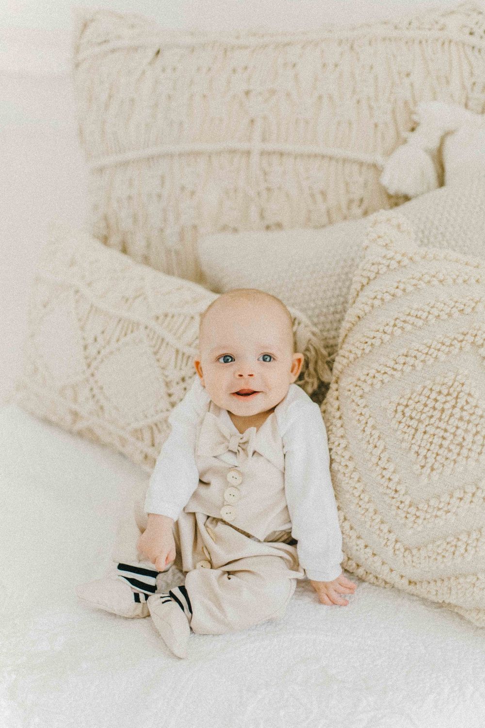 Baby wearing beige outfit sits among cream-colored textured pillows on white couch. Fotografia profissional de maternidade, recém-nascido.