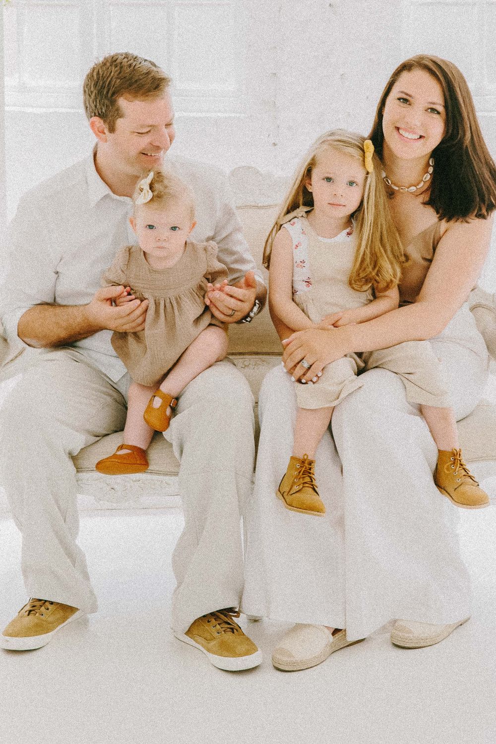 Family dressed in light neutral colors sits together on white couch.