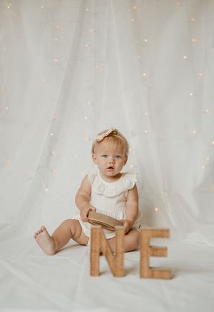 Small child in white dress sitting on white backdrop with wooden letters spelling ONE and twinkling lights.