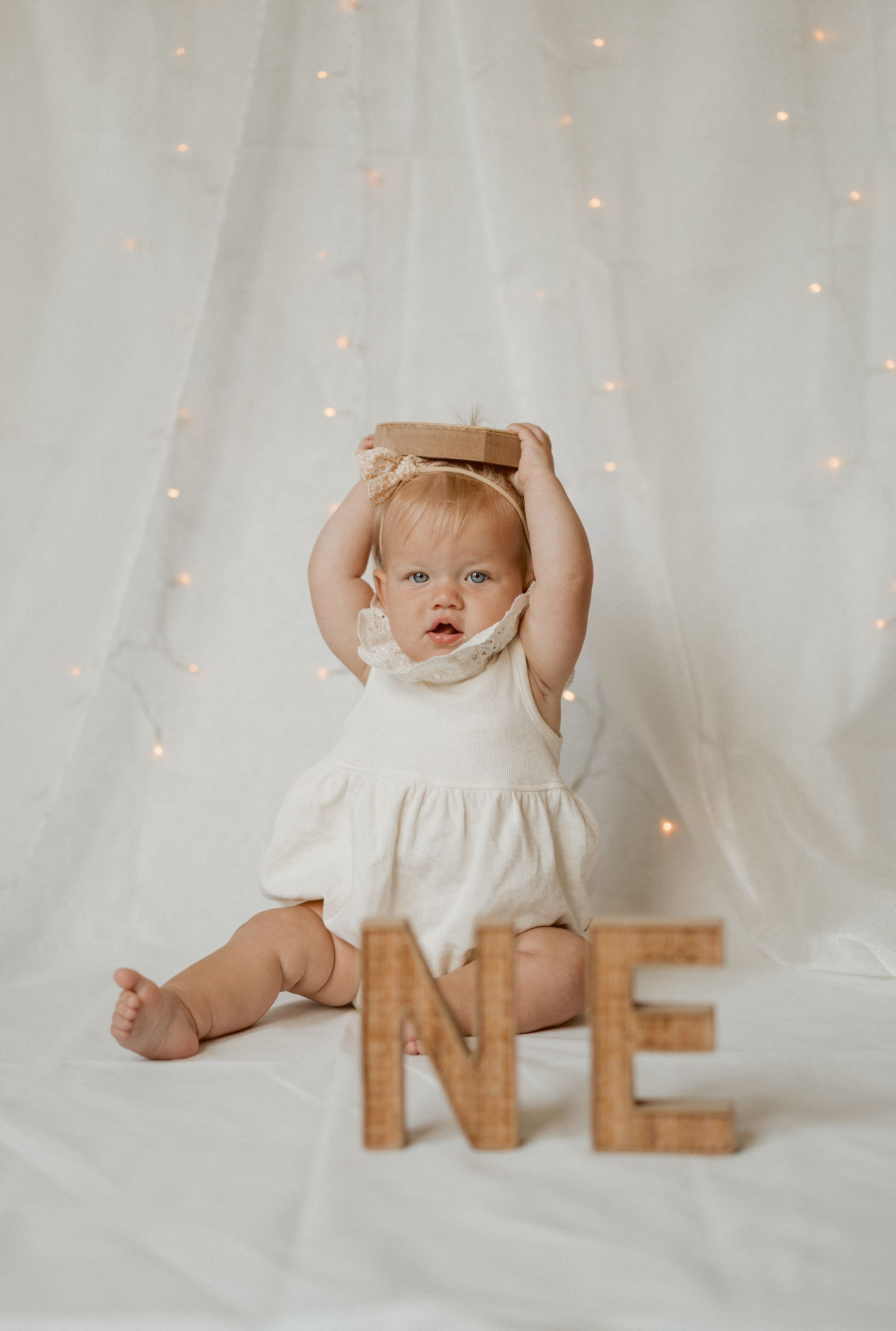 Baby in white dress poses with arms raised near wooden ONE letters against sparkly backdrop.