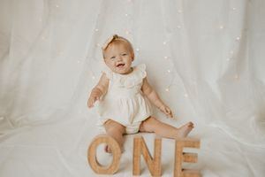 Baby in white outfit sits on fabric backdrop with wooden letters spelling 'ONE' for first birthday photo shoot.